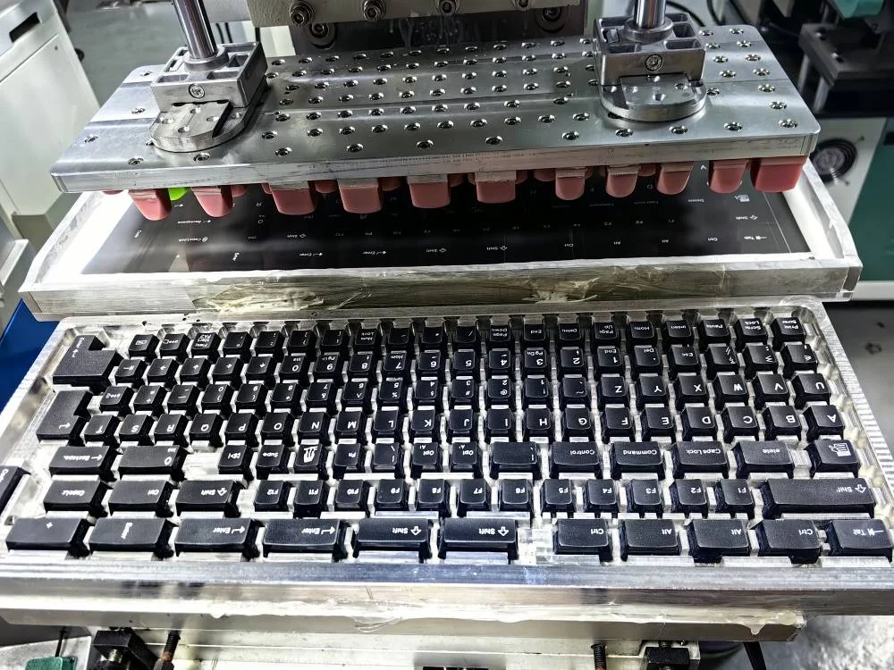 As of late May, here is the latest factory photo showing a test print. This is not yet approved for production. They still need to fine tune the alignment to make it more consistent; a few keys on one side look to have been double printed and a number keys did not have full contact, such as tab. They said that the machine they recommended did not have enough pressure for pad printing the new Model F keys, so they are spending a couple weeks to build another machine that has higher pressure capability. The custom equipment pictured below was the big time commitment whereas the actual machine is standardized so they just need to switch the custom tooling to the new machine instead of needing to do all of the custom stuff again. They said they expect to wrap up the pad print process around month end!